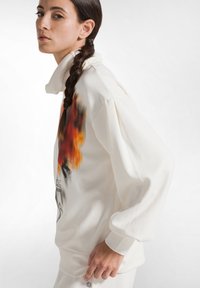 Mujer con el cabello trenzado, vistiendo un sudadera blanca con un diseño de llama colorido, de pie contra un fondo blanco sencillo.