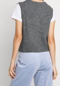 Top senza maniche grigio in maglia con una texture a coste, indossato sopra una t-shirt bianca, abbinato a pantaloni in velluto a coste di colore blu chiaro.