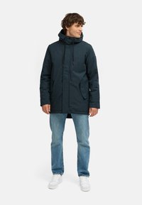 Tmavě modrý parka kabát s kapucí, zipovým uzávěrem, nastavitelnými šňůrkami, dvěma bočními kapsami a středně dlouhým střihem, spárovaný se světlými džíny.