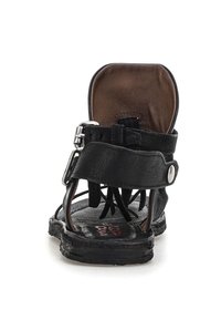 Sandal de cuero negro con correa de hebilla, área del talón acolchada en marrón y flecos decorativos en la parte delantera de la suela, visto desde atrás.