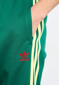 Gröna träningsbyxor med gula sidostråk, med en röd broderad Adidas-logga på vänster lår och en sidficka.