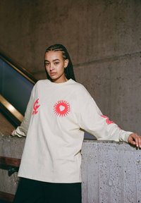 Lichtbeige langslijstshirt met rode grafische ontwerpen op de voorkant en mouwen. Ontspannen pasvorm, zachte textuur en ronde hals.