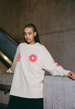 Camisa de manga larga color beige claro con diseños gráficos rojos en la parte frontal y las mangas. Corte relajado, textura suave y escote tipo crew.