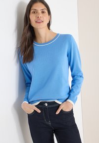 Pull en tricot bleu avec col rond, poignets et ourlet côtelés. Associé à un jean foncé avec des coutures blanches et un bouton à l'avant.