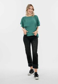 Blusa turchese con maniche a sbuffo, fianchi arricciati, abbinata a jeans scuri arrotolati sui polsini e sneaker nere. Texture liscia.