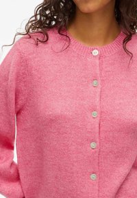 Femme portant un cardigan en tricot rose avec cinq boutons blancs et des cheveux bruns bouclés visibles au niveau du col.