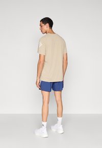 A bézs rövid ujjú póló jobb vállán három fehér csíkkal, kék rövidnadrággal és fehér tornacipővel párosítva.