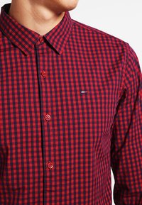 Man met een rood en marineblauw geblokt overhemd met een klein geborduurd logo op de borst.
