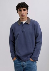Long-sleeve navy polo shirt with a contrasting brown corduroy collar, two-button placket, and side slits, paired with light denim jeans.
