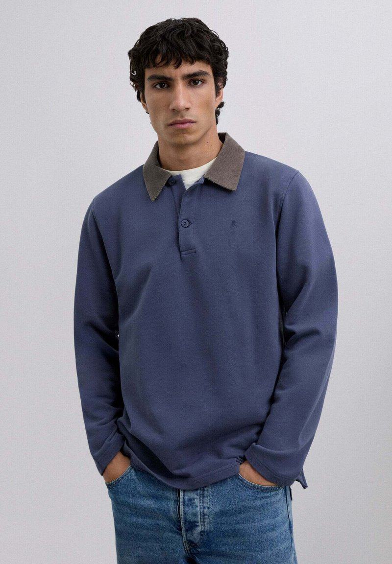 Long-sleeve navy polo shirt with a contrasting brown corduroy collar, two-button placket, and side slits, paired with light denim jeans.