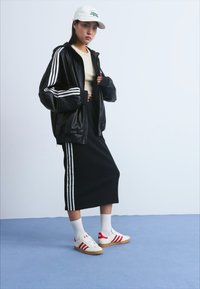 Juodas zip-up džemperis su baltais dryžiais, šviesiai rudas viršus, juodos sijonas su šoninėmis juostelėmis, baltos kojinės, raudoni ir balti sportbačiai, bei balta kepurė.