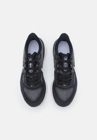 Des chaussures de sport noires avec une tige en mesh présentent un design texturé, un bout rond et un logo blanc contrastant. Les lacets sont noirs et la languette est rembourrée.