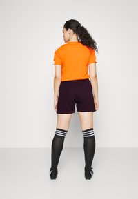 Kurze ärmellose orange Trikot, schwarze Shorts, schwarze Kniestrümpfe mit weißen Streifen und schwarze Fußballschuhe. Rückansicht des aktiven Outfits.