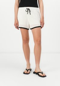 White woven shorts with black scalloped trim, elastic waistband, and drawstring. Paired with black flip-flops and a black top.