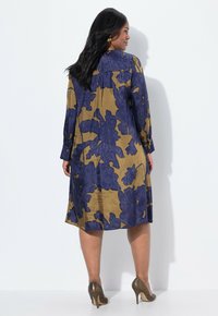Vestido envolvente floral en mostaza y azul marino. Mangas largas, diseño fluido, textura suave. Presenta pliegues sutiles y un dobladillo a la altura de la rodilla.