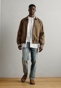 Brown bomber jacket with elastic cuffs, white shirt underneath, light blue jeans, and brown shoes on wooden floor. Neutral background.