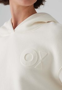 Crème-kleurige hoodie met een trekkoordkap, voorzien van een verdiept rond en driehoekig logo op de voorkant. Zacht, textuurstof.