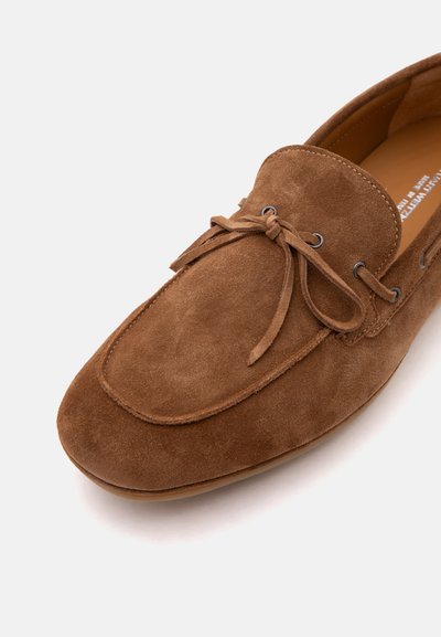 Mocassin en daim marron avec un nœud sur le dessus, présentant des détails cousus et des œillets métalliques sur fond blanc.