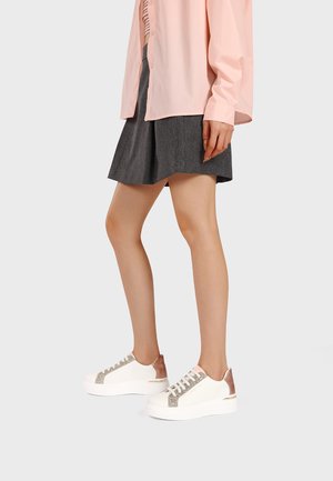 Donna che indossa sneakers con zeppa bianche con glitter e accenti bronzo, abbinate a una gonna grigio scuro e una camicetta rosa chiaro.