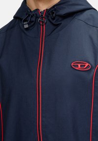 Granatowa kurtka z czerwonymi akcentami, z zamkiem błyskawicznym, regulowanym kapturem i logo. Gładka tekstura i lekki materiał.
