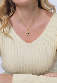 Goldenes Halsband mit einem herzförmigen Anhänger, der mit klaren Kristallen verziert ist. Das Model trägt ein cremefarbenes, geripptes Oberteil mit einem V-Ausschnitt.