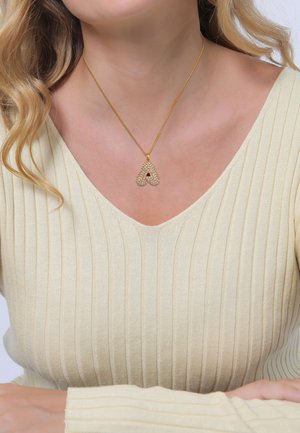 Goldenes Halsband mit einem herzförmigen Anhänger, der mit klaren Kristallen verziert ist. Das Model trägt ein cremefarbenes, geripptes Oberteil mit einem V-Ausschnitt.
