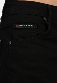 Jeans in denim nero con una tasca posteriore decorata con un'etichetta cucita rossa e bianca "CIPO & BAXX". Presentano dettagli come rivetti in metallo e una texture liscia.