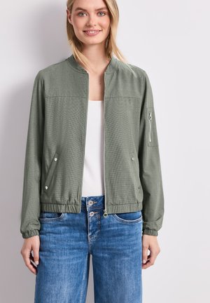 Lächelnde Frau trägt eine olivgrüne Bomberjacke mit Reißverschluss über einem weißen Top und blauen Jeans, steht vor einem schlichten hellen Hintergrund.