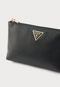 Pochette synthétique noire à surface texturée, ornée d'une plaque logo triangulaire dorée et d'une fermeture éclair supérieure. Design compact et élégant.