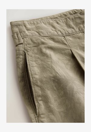Khaki pleated shorts made of textured fabric, featuring side pockets and stitching detailing. The surface shows a subtle floral pattern.