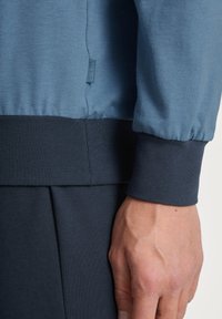 Blauwe sweatshirt met geribbelde marineblauwe manchetten en zoom. Gladde textuur, casual ontwerp. Etiket zichtbaar op de zijnaad. Hand rustend tegen de stof.