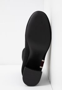 Semelle d'une botte noire à talon avec un bout arrondi et un talon carré, reposant contre un angle de mur et de sol blancs.