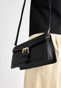 Czarna skórzana torebka na ramię o wyraźnym kształcie, z złotymi akcentami, przednim klapą i plecionym paskiem, ozdobiona detalem w postaci zapięcia.