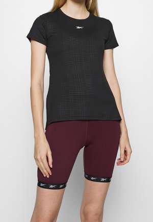 Kvinde iført en sort kortærmet top med et diskret ternet mønster og logo, kombineret med bordeaux cykelshorts med sorte logo-bånd ved kanten.