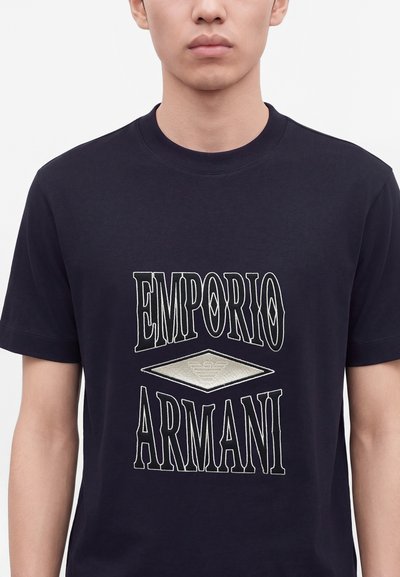 Νέος άνθρωπος φοράει μαύρο κοντομάνικο πουκάμισο με μεγάλο λευκό περίγραμμα κειμένου που γράφει Emporio Armani και ασημένιο διαμάντι λογότυπο στο κέντρο.