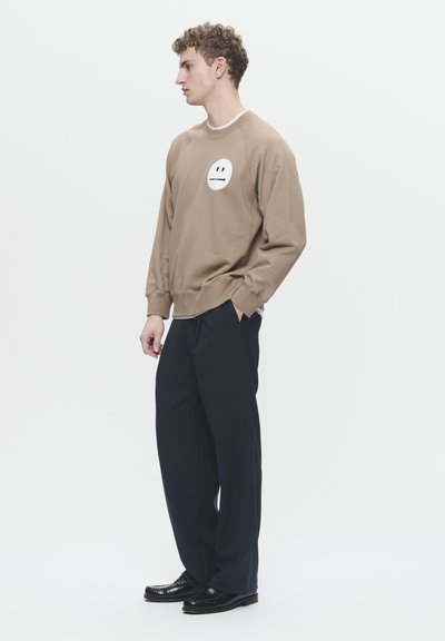 Felpa marrone con una grafica rotonda bianca che presenta un design di faccia minimalista, abbinata a pantaloni navy scuri e scarpe nere.