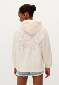 Crème hoodie met een achterzijde design van een schedel en gekruiste botten in roze studdetails. Gemaakt van zachte katoenen stof, met een relaxte pasvorm en een kangoeroezak.