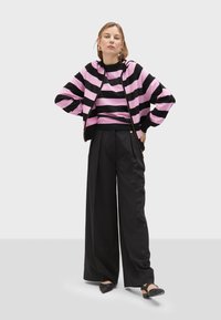 Felpa con zip a strisce rosa e nere sopra una maglietta nera, abbinata a pantaloni neri a gamba larga e scarpe nere basse. Gli orecchini dorati sono evidenti.