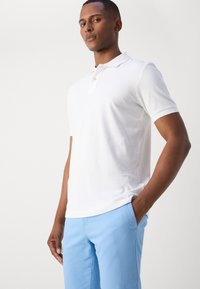 Hvid polo skjorte med krave, der har korte ærmer og et diskret logo. Båret med lyseblå bukser, stoffet ser glat og afslappet ud.