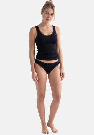 Donna con capelli biondi raccolti in uno chignon, che indossa un top nero con bordi in pizzo e biancheria intima nera coordinata con bordi in pizzo, in piedi a piedi nudi su sfondo bianco.