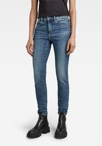 Jeans de mezclilla azul de corte ajustado, con un acabado desgastado, diseño clásico de cinco bolsillos y cierre de botón, combinados con botas negras de tobillo grueso.