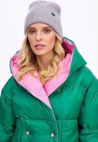 Zielona pikowana kurtka z różową podszewką i zatrzaskami, zestawiona z jasnoszarym dzianym beanie z małym zielonym akcentem.