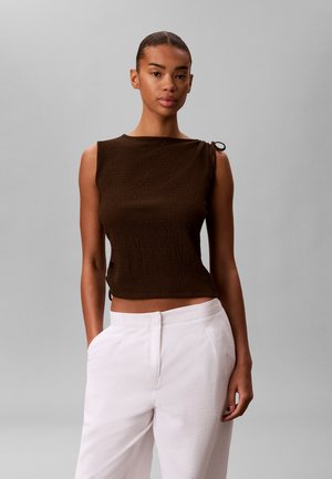 Vrouw draagt een mouwloze donkerbruine gestructureerde top met een klein schouderstrikje, gecombineerd met een witte broek met hoge taille, staand tegen een grijze achtergrond.