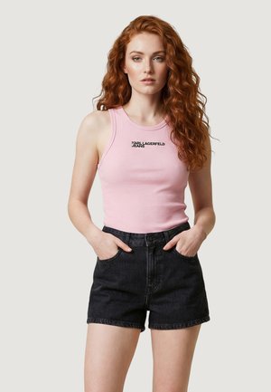Joven mujer con cabello largo, rojo y rizado lleva un top rosa sin mangas con "Karl Lagerfeld Jeans" y shorts vaqueros negros, con las manos en los bolsillos.