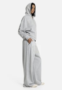 Γυναίκα που φοράει ανοιχτό γκρι φαρδιά φούτερ με διακοσμητικά καρφιά και αντίστοιχο φαρδύ παντελόνι φόρμας, στέκεται με το ένα χέρι στην τσέπη.