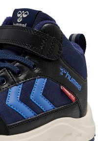 Nahaufnahme eines navyblauen und schwarzen Sneakers mit blauen Chevron-Streifen, schwarzem Riemen und weißer Sohle, auf dem das Hummel-Logo und das Climatex-Tag sichtbar sind.