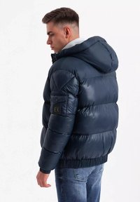 Giubbotto puffer blu navy con cappuccio, texture lucida e polsini elastici. Presenta una patch sul braccio sinistro e una vita aderente. Jeans visibili sotto.