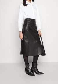 Gonna midi in pelle nera con taglio dritto, caratterizzata da bottoni laterali. Abbinata con una blusa bianca e stivaletti neri con tacco. Borbono nero incluso.