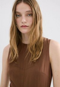 Bruin mouwloos topje met een hoge halslijn, een gladde textuur en subtiele stikdetails aan de voorkant; model met lang, golvend haar.