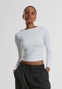 Weißes langärmliges Crop-Top mit Bootsausschnitt und Raffung, kombiniert mit hoch taillierten schwarzen Streifenhosen. Glatte Stoffstruktur.